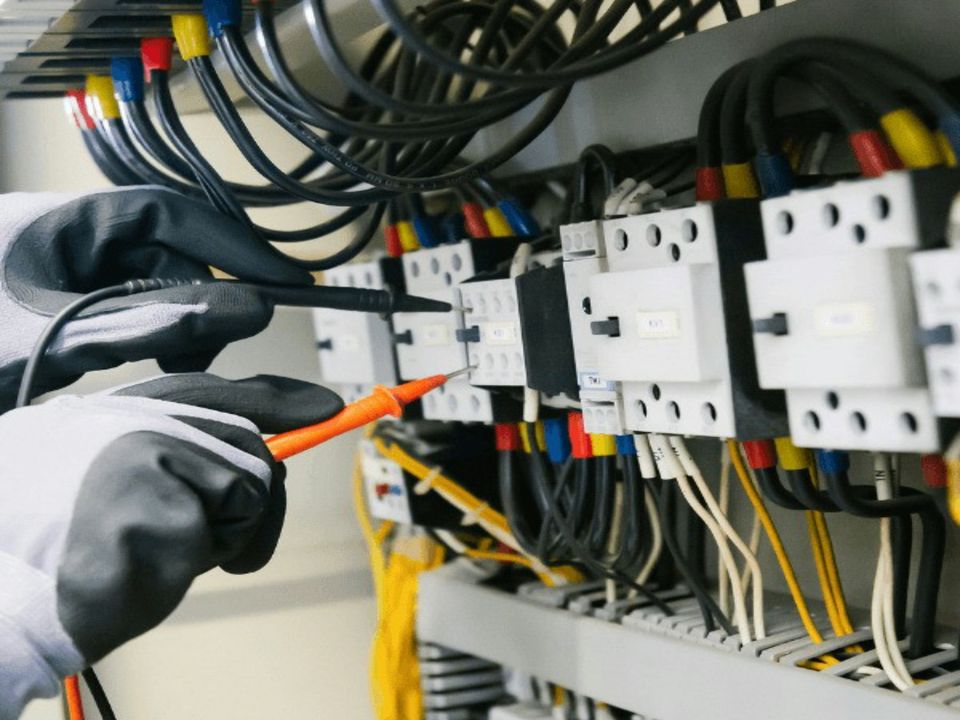 Gloved hands using a multimeter to test terminals inside an electrical panel