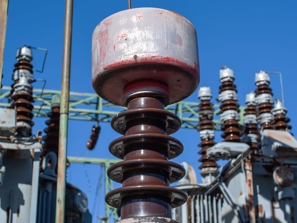 Close-up of a high-voltage insulator stack at a substation, with transformers in the background